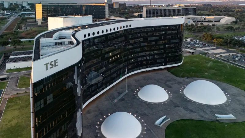 Vista aérea de um prédio corporativo com fachada branca e a sigla “TSE”, cercado por áreas verdes e iluminação no pátio com três estruturas circulares no telhado.