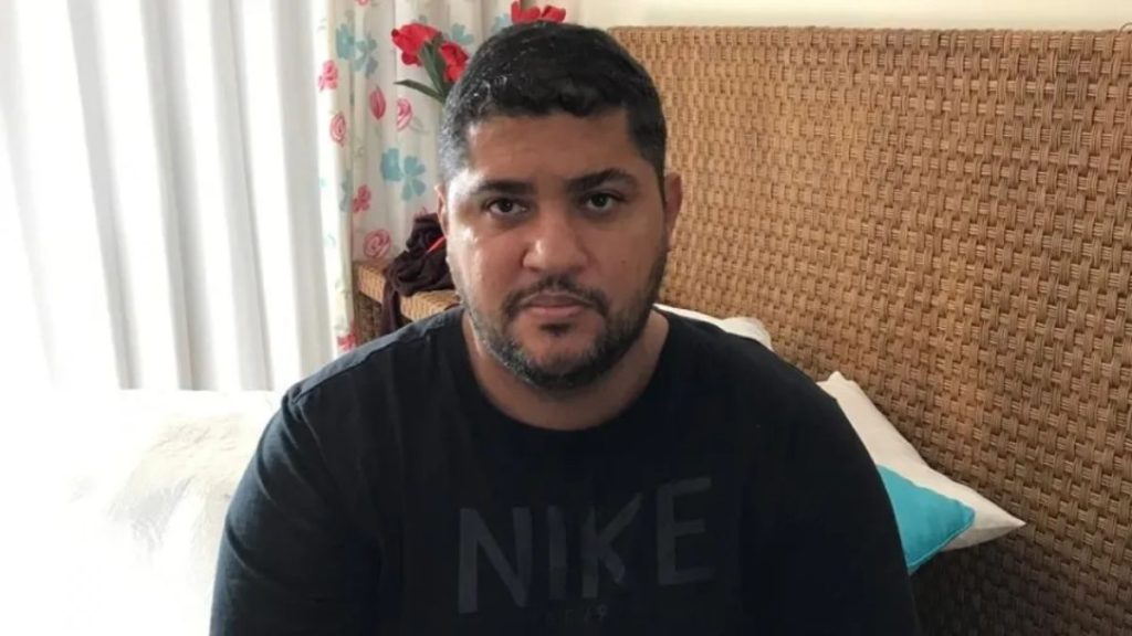 Homem de barba e cabelo escuros, usando camiseta preta, em ambiente interno sentado em uma poltrona de tecido, em foto de reprodução relacionada a “Homem ligado a André do Rap é preso”.