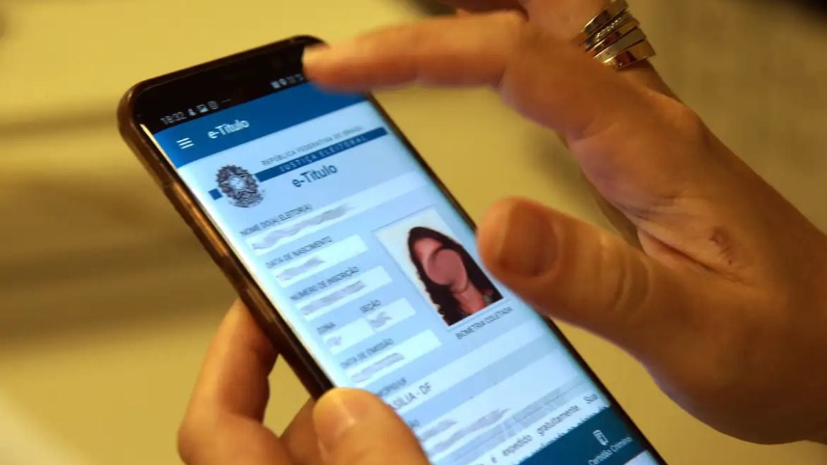 Mão segurando um celular com a tela do e-Título aberta, mostrando informações para baixar o e-Título e usar na votação, em ambiente interno.