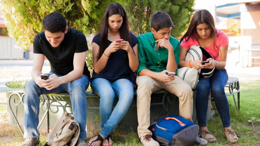 Quatro jovens sentados no gramado ao lado de uma cerca viva, cada um usando o celular e olhando para as telas enquanto descansam e conversam.