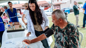 Voluntária ajuda homem sentado em evento ao ar livre, realizando atendimento com materiais sobre a mesa e garantindo suporte durante a ação comunitária.