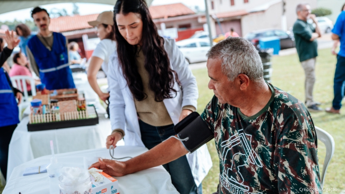 Voluntária ajuda homem sentado em evento ao ar livre, realizando atendimento com materiais sobre a mesa e garantindo suporte durante a ação comunitária.