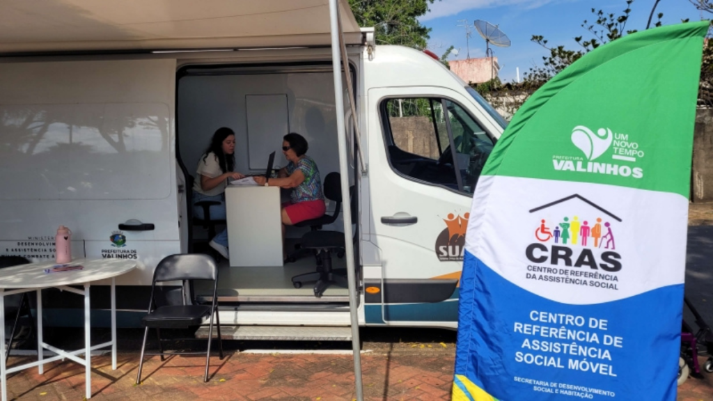 Equipe realiza atendimento em ação social dentro de uma van branca, com banner do Centro de Referência de Assistência Social (CRAS) e Centro de Referência em Assistência Social Móvel na praça