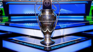 Taça da Champions League em destaque sobre mesa em um estúdio de transmissão, com fundo azul e telas, indicando cobertura do sorteio da fase de Liga