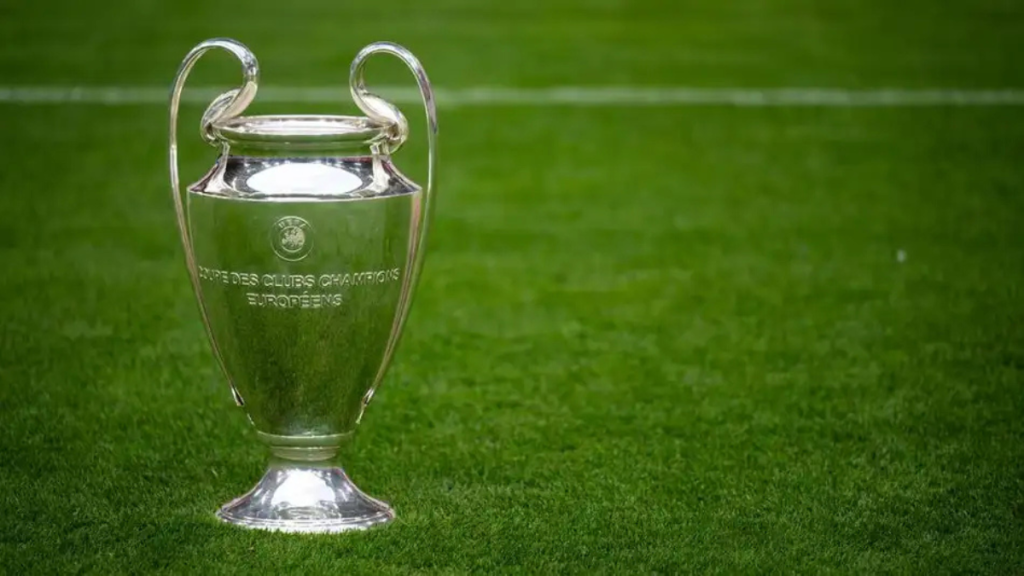 Troféu da Champions League sobre o gramado verde, em destaque, com espaço ao lado para mostrar como ficou o sorteio da fase de Liga da Champions League 2026/27.