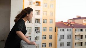 regras de condomínio: Mulher em varandas de um prédio de apartamentos observa o condomínio, em contexto de esclarecimentos do síndico sobre barulho e gás; foco em regras de convivência.