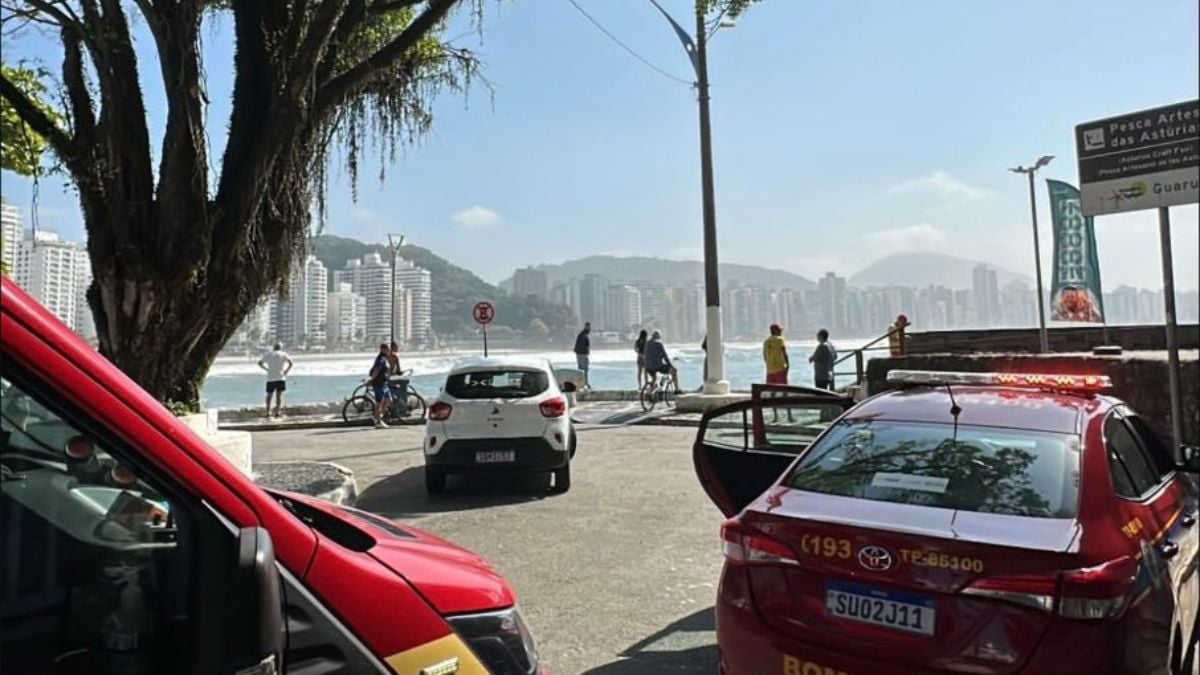Afogamento na Praia das Astúrias em Guarujá: Cena em via à beira-mar na Praia das Astúrias, em Guarujá, com carros estacionados e agentes na área durante ocorrência envolvendo afogamento, com pessoas ao fundo observando a orla.