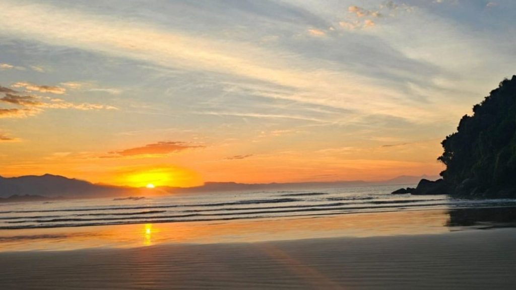 Praia de Itaguaré, Bertioga: praia com mar calmo, reflexo do sol na areia, ondas suaves e vegetação da Mata Atlântica ao lado do litoral.