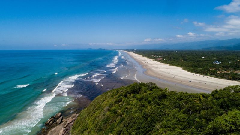 Guaratuba, vista aérea de praia com mar azul e faixa de areia ao lado de vegetação da Mata Atlântica e estrada costeira.