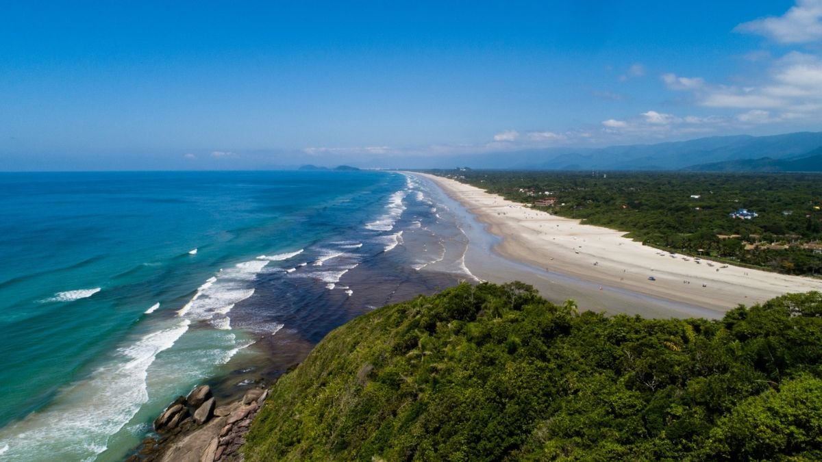 Guaratuba, vista aérea de praia com mar azul e faixa de areia ao lado de vegetação da Mata Atlântica e estrada costeira.