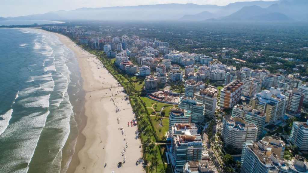 Riviera de São Lourenço: Vista aérea da orla de Guaratuba com praia extensa ao lado da cidade, prédios e faixa verde, mostrando a experiência entre mar, rio e Mata Atlântica