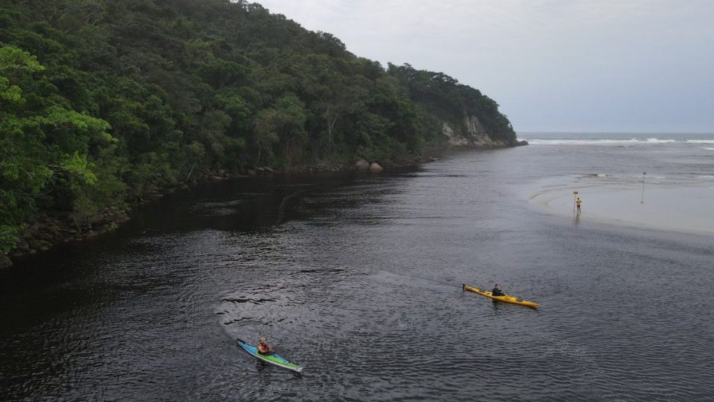 Guaratuba com paisagem costeira entre o mar e a Mata Atlântica, com caiaques navegando em águas calmas ao longo da praia e da mata verde na encosta