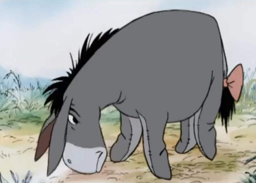 Burro Bisonho, de Ursinho Pooh, aparece em uma cena na floresta, caminhando pelo caminho com expressão triste. Peter Cullen dublou o personagem.