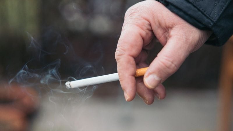 Dia Nacional de Combate ao Fumo: pessoa segurando um cigarro entre os dedos, com fumaça visível, lembrando sinais de alerta na boca, olhos e voz que fumantes não devem ignorar