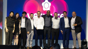 Grupo de pessoas em foto de premiação, com oito participantes em pé no palco, ao lado de um vencedor com certificado e troféu, diante de telão com logotipo colorido e iluminação rosa e azul.
