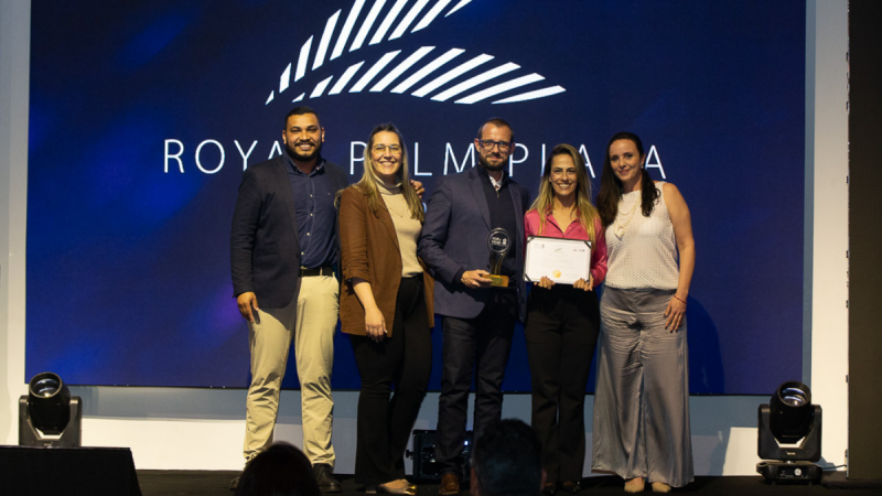 Grupo de pessoas posando no palco durante evento com backdrop azul escrito “ROYAL PALM PLAZA”, incluindo uma pessoa recebendo um certificado. Foto institucional.