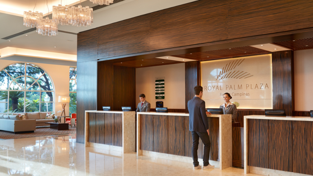 Recepção de hotel com equipe atendendo em balcões de madeira, luzes modernas no saguão e painel com o texto “ROYAL PALM PLAZA” e “Rio Campinas”.