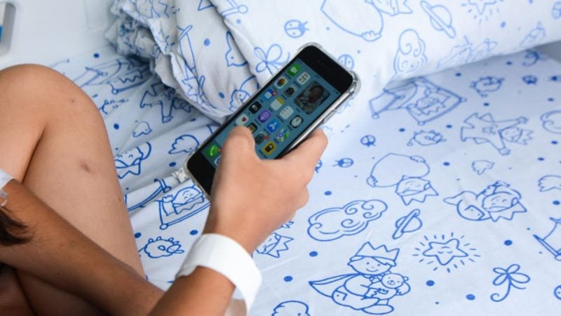 Criança usando smartphone na cama, com ilustração de desenhos no lençol, sugerindo discussão sobre uso excessivo de telas e TDAH em crianças.