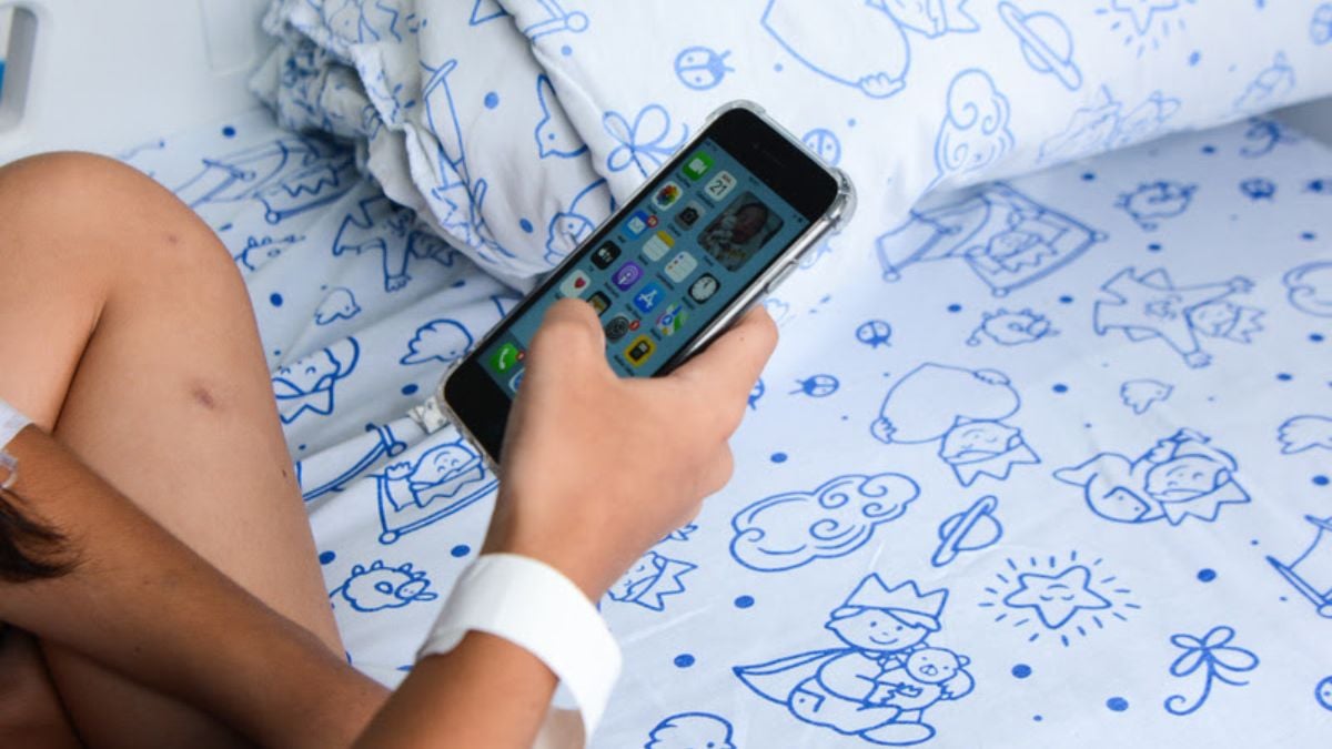 Criança usando smartphone na cama, com ilustração de desenhos no lençol, sugerindo discussão sobre uso excessivo de telas e TDAH em crianças.