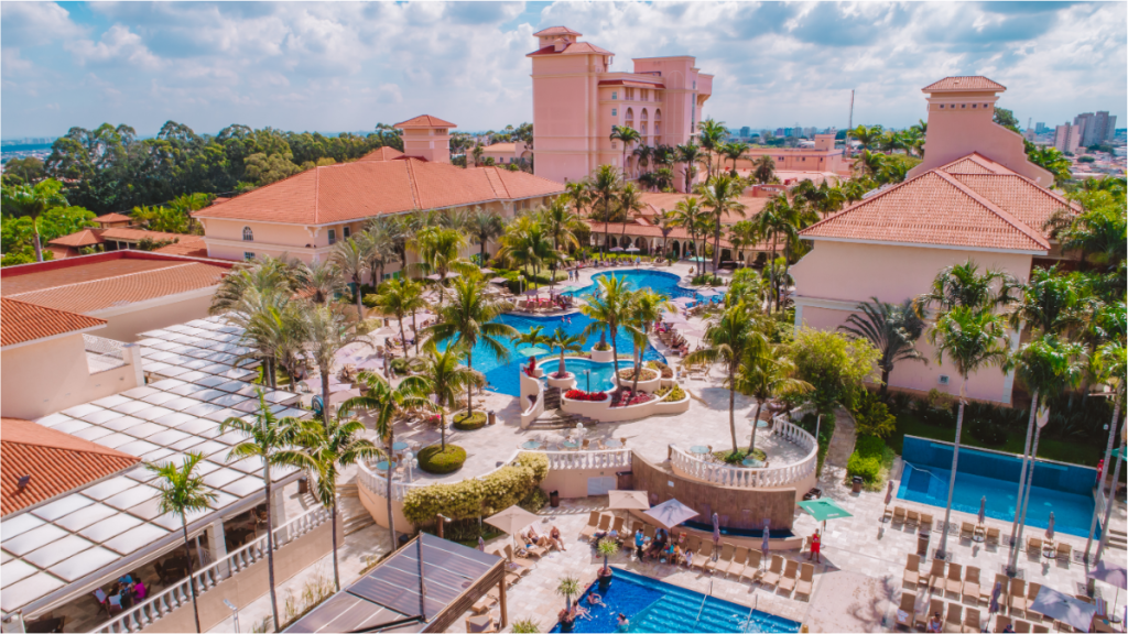 Vista aérea Royal Palm Plaza com piscinas azuis, áreas de lazer, jardins com palmeiras e prédios com telhados de cerâmica, em meio a vegetação.