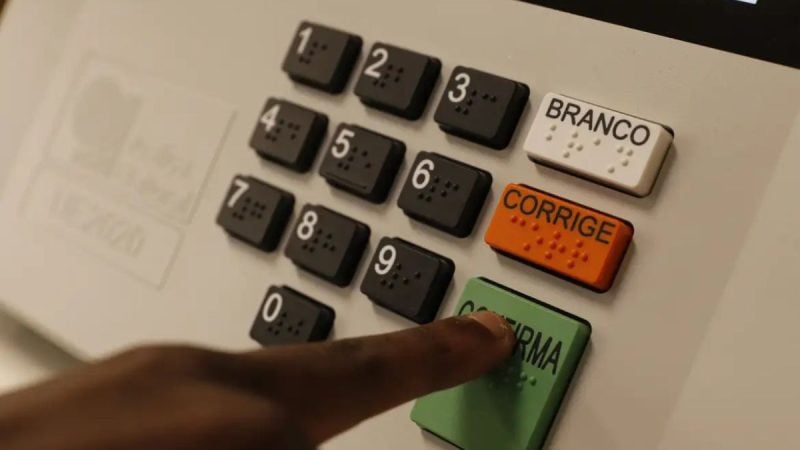 Imagem de uma urna eletrônica em eleições 2026 com botões numerados e as opções BRANCO e CORRIGE; uma mão aponta para o botão de uma candidatura, mostrando como votar e usar a cola eleitoral