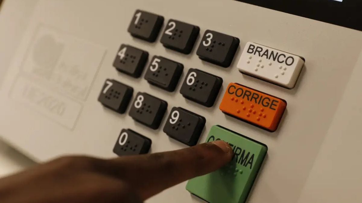 Imagem de uma urna eletrônica em eleições 2026 com botões numerados e as opções BRANCO e CORRIGE; uma mão aponta para o botão de uma candidatura, mostrando como votar e usar a cola eleitoral