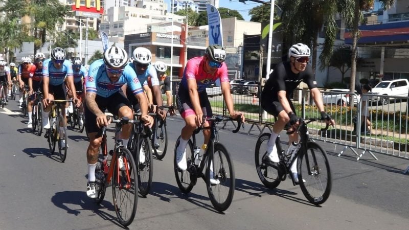 Ciclistas em competição pedalam em grupo em uma avenida durante um evento esportivo ao ar livre, com capacetes e bicicletas alinhadas na pista.
