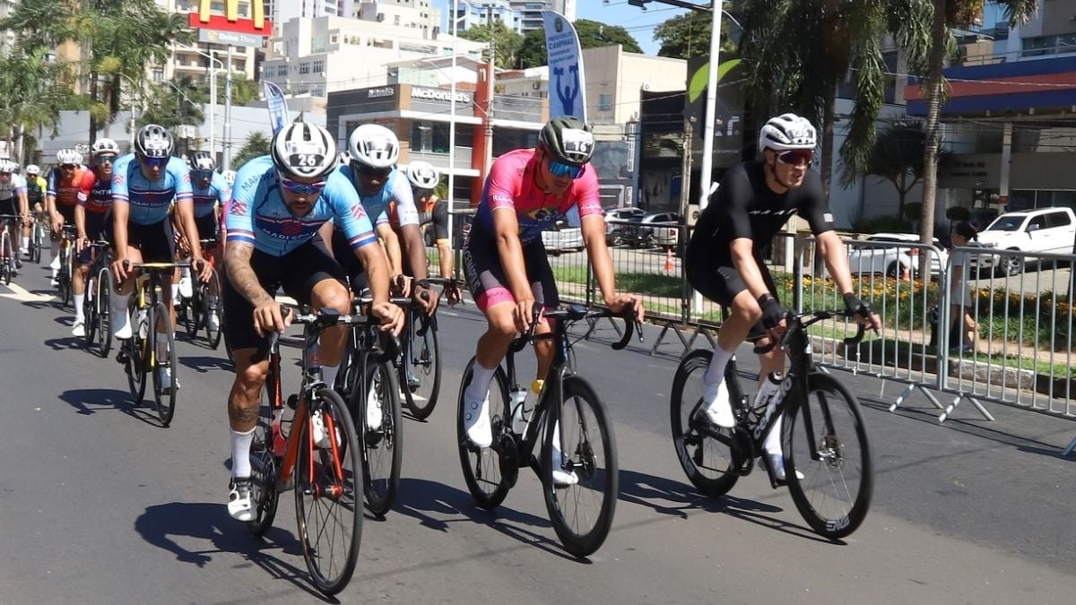 Ciclistas em competição pedalam em grupo em uma avenida durante um evento esportivo ao ar livre, com capacetes e bicicletas alinhadas na pista.