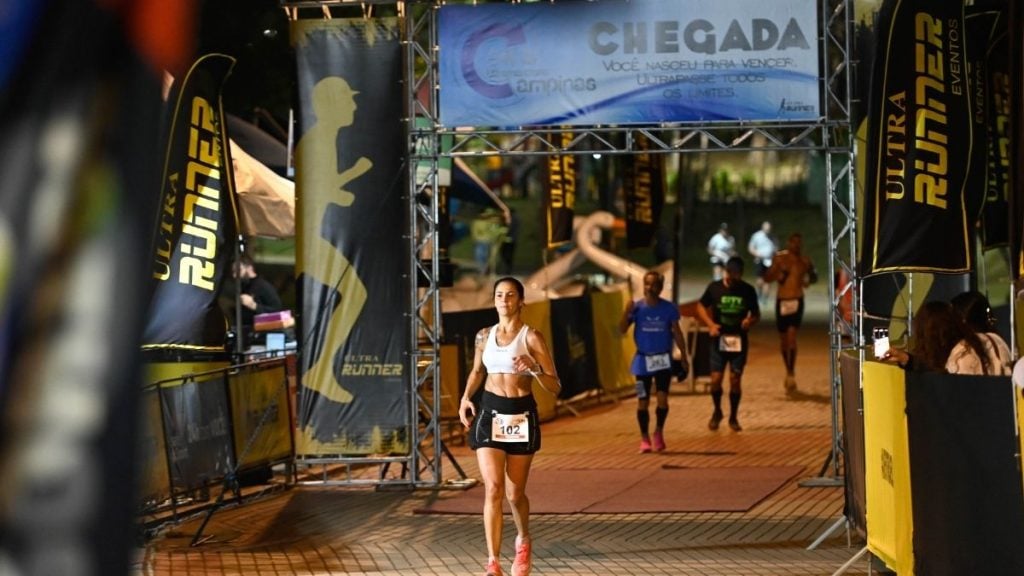 Cena de chegada em prova de corrida: uma atleta passa pelo pórtico de largada com número 102, enquanto outros participantes correm ao fundo em ambiente iluminado.