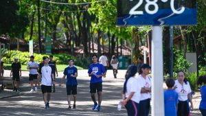 Pessoas caminhando em uma área arborizada ao ar livre, em um dia ensolarado, com placas de orientação na entrada do caminho e estudantes seguindo pela rua.
