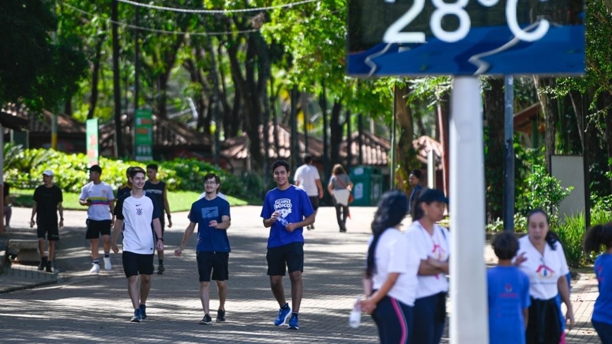 Pessoas caminhando em uma área arborizada ao ar livre, em um dia ensolarado, com placas de orientação na entrada do caminho e estudantes seguindo pela rua.
