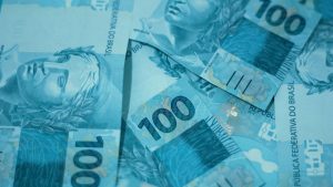 10 maiores bilionários do Brasil — Forbes divulga o ranking com uma imagem de cédulas de dinheiro brasileiro em tom azul, destacando notas de 100 reais e 10 reais ao fundo.