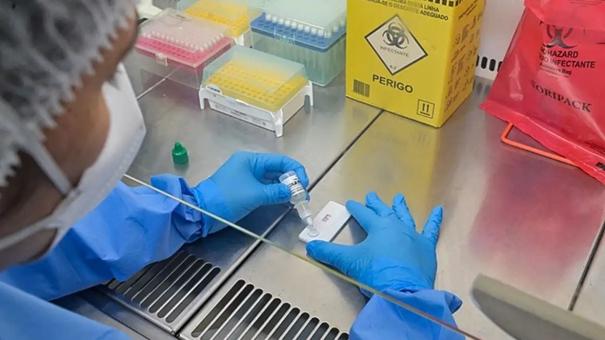 Técnico em laboratório usando luvas azuis e equipamento automatizado para manipular uma amostra em tubo, ao lado de materiais descartáveis e caixas de reagentes de risco.