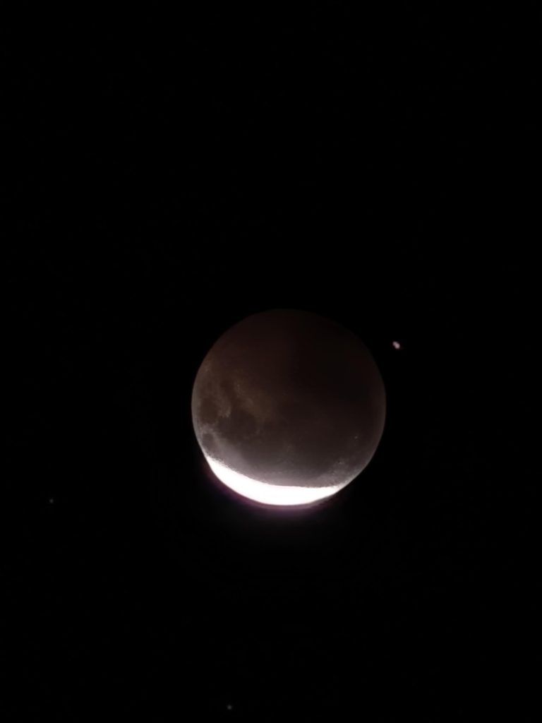 Lua quase cheia vista no céu escuro, com parte iluminada e sombra formando um disco avermelhado ao centro. Brilho intenso na borda inferior.