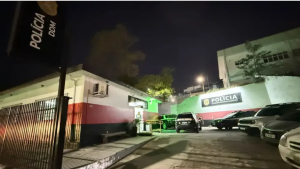 Cena noturna em frente a um prédio público com viaturas e policiais ao lado, iluminada por luzes externas e placas da polícia, em registro sobre importunação sexual em Tuiuti