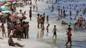 Banhistas lotam uma praia na Baixada Santista durante onda de calor, com pessoas se refrescando no mar e outros sob guarda-sóis e cadeiras; clima de até 35°C.