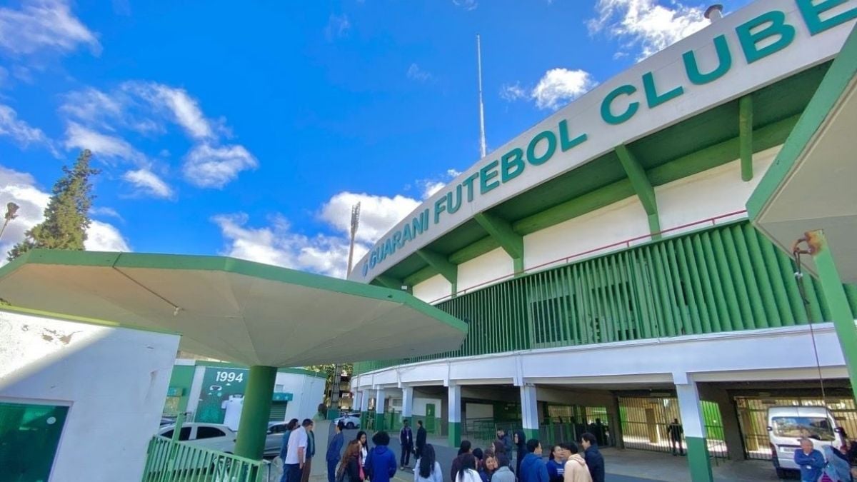 Vista externa do estádio do Guarani, com arquibancada e fachada verde do clube, pessoas na entrada.