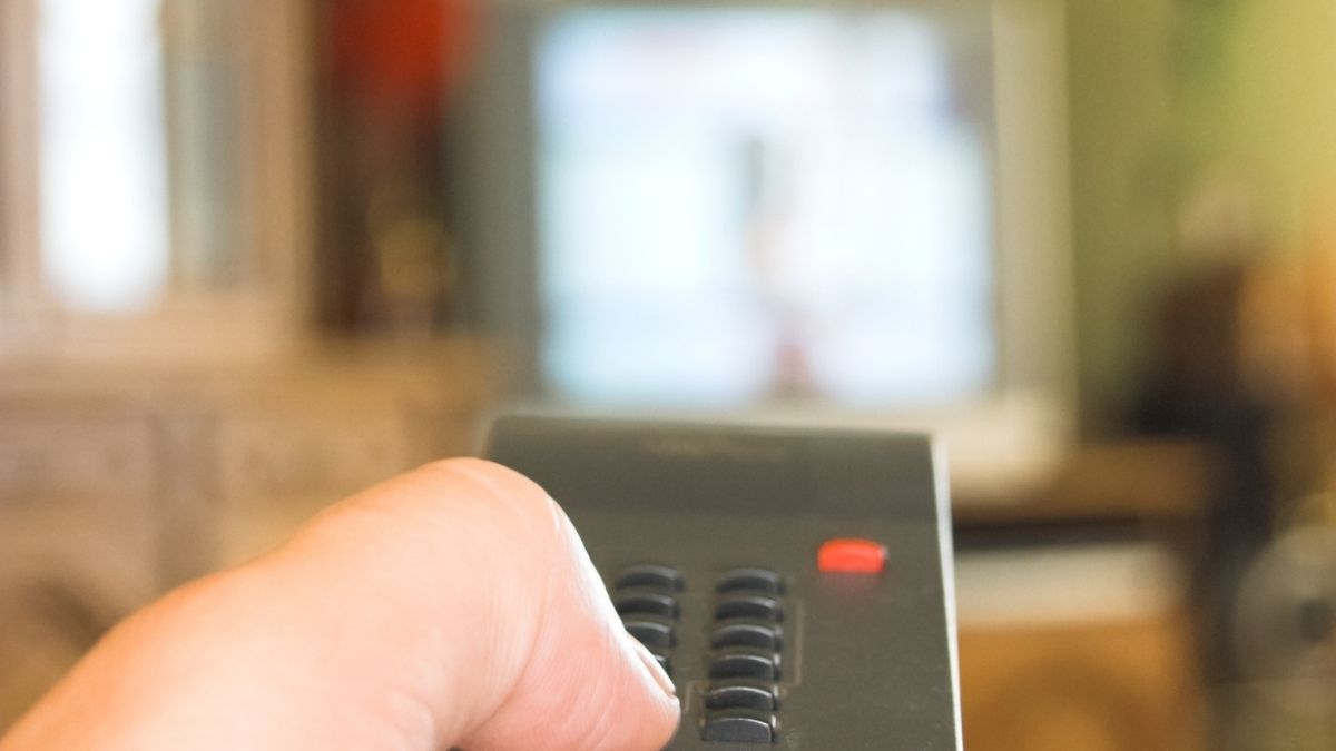 Mão segurando um dispositivo eletrônico preto com botões e luz indicadora laranja, em ambiente interno desfocado, sugerindo controle ou acionamento de equipamentos no trabalho.