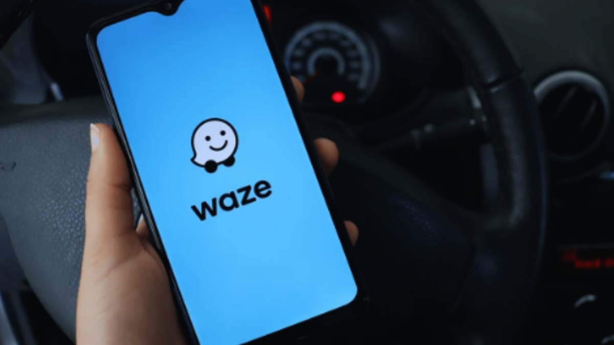 Pessoa segurando um celular com o aplicativo Waze aberto na tela, mostrando o logo do Waze em fundo azul, enquanto ao fundo há o painel de um carro.