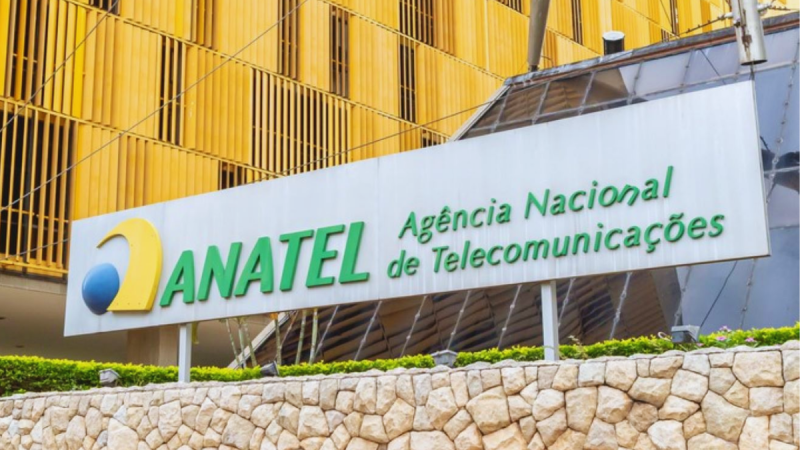 Placa de fachada da ANATEL e o texto “Agência Nacional de Telecomunicações”, em frente a um prédio com estruturas amarelas e jardim, no Brasil.