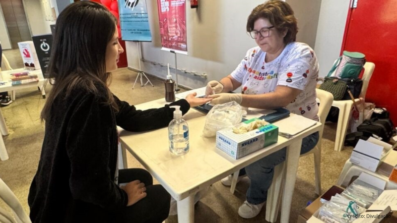 Duas pessoas conversam em uma mesa durante atendimento, com profissional usando jaleco e óculos ao lado de itens de cuidado e materiais organizados em ambiente interno.