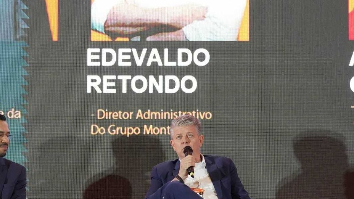 Palestra com o executivo Edovaldo Retondo sentado em uma cadeira, segurando um microfone, em evento corporativo no palco; ao fundo há telão com a inscrição “Painelista”.