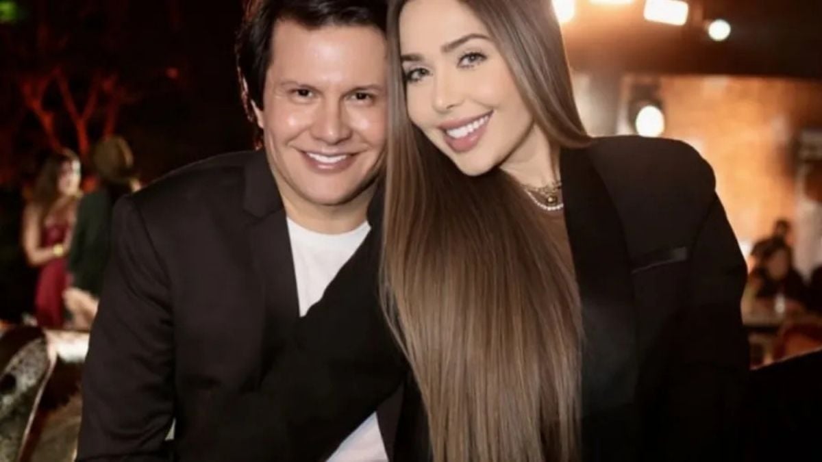 Foto de casal em evento social, com um homem de terno escuro ao lado de uma mulher com cabelo longo e vestido escuro, ambos sorrindo em ambiente iluminado ao fundo.
