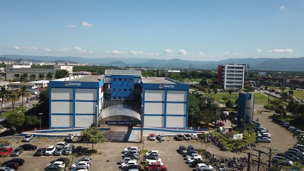 Vista aérea de um prédio industrial azul e branco em Guarujá, com estacionamento ao redor, carros estacionados e paisagem urbana e montanhas ao fundo.