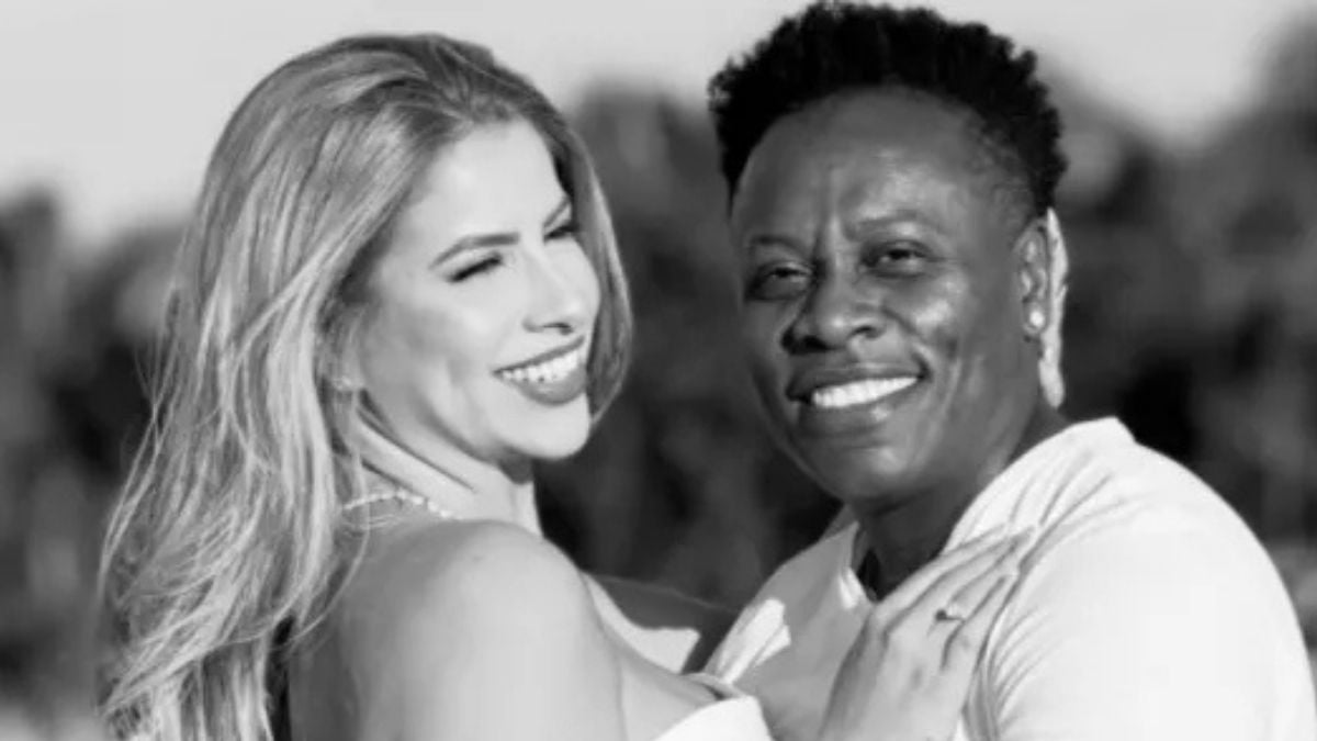 Casal sorridente em preto e branco, a mulher abraça o parceiro em um cenário externo com folhagens desfocadas ao fundo.