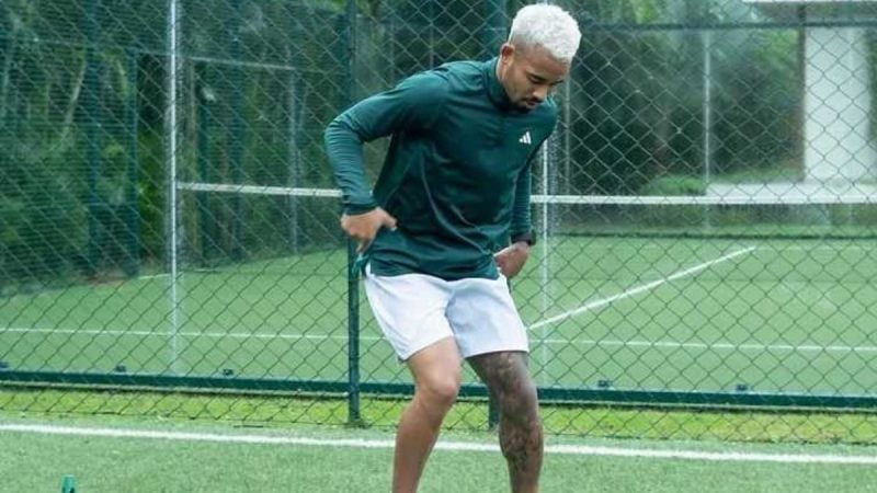 Homem em treino de futebol em quadra com rede: ele usa uniforme verde e branco, faz controle de bola e aparece inclinado durante o exercício.