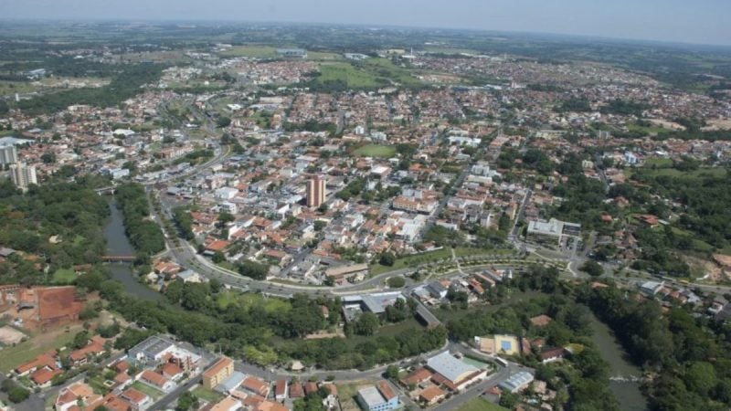 Vista aérea de uma cidade brasileira com ruas e bairros residenciais, telhados de casas, áreas verdes e um rio serpenteando pelo centro, em dia claro.