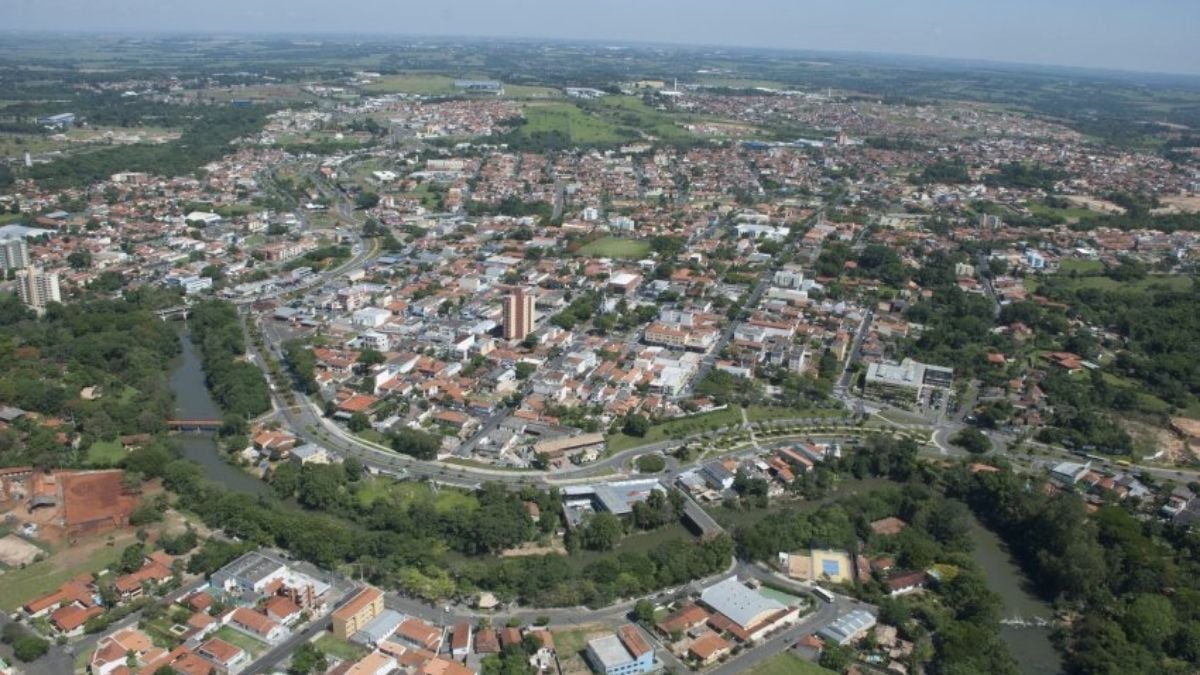 Vista aérea de uma cidade brasileira com ruas e bairros residenciais, telhados de casas, áreas verdes e um rio serpenteando pelo centro, em dia claro.