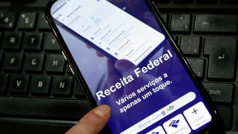 Celular mostrando aplicativo com a mensagem “Receita Federal” e referência a variações, serviços e toque, em fundo de teclado de computador, com contexto de restituição do Imposto de Renda.