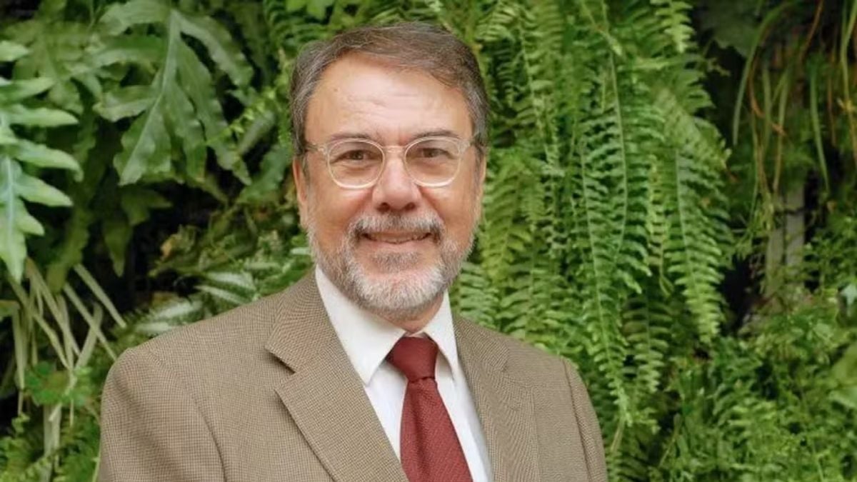 Homem adulto com cabelo grisalho e barba, usando óculos e terno bege com camisa branca e gravata vermelha, posando diante de um fundo com plantas verdes.